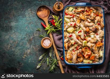 Baked Chicken with vegetables in casserole with wooden spoon and fresh herbs and spices on dark rustic background, top view