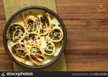 Baked crepe roll casserole filled with spinach, tomato and mincemeat with cream sauce and grated cheese on top, served in rustic bowl, photographed overhead on dark wood with natural light (Selective Focus, Focus on the top of the food)