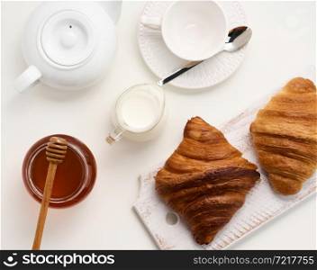 baked croissants, white ceramic teapot and empty cup and saucer, jar of honey on a white table, top view. Breakfast
