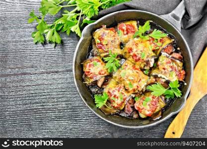 Baked eggplant rolls with meat, tomatoes and cheese in a frying pan, napkin, parsley and spatula on a dark wooden board background from above