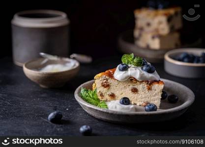 Baked homemade cottage cheese casserole or pudding with raisins serving with blueberry and sour cream on dark background. Close-up.. Cottage cheese casserole