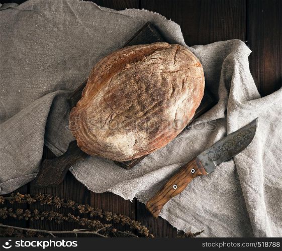 baked oval bread made from rye flour on a wooden cutting board, knife lies next to it, top view