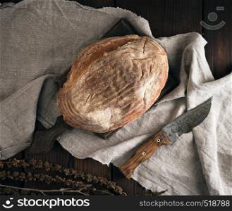 baked oval bread made from rye flour on a wooden cutting board, knife lies next to it, top view