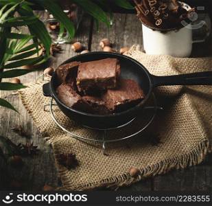 baked pieces of chocolate brownie cake with walnuts in a black metal frying pan on a wooden table, top view