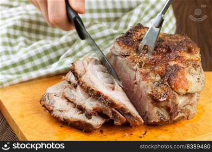 Baked pork meat with spices sliced on brown cutting board with knife and fork. Grilled meat. Man cuts piece of meatn on slices with knife.. Baked pork meat with spices on wooden cutting board