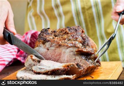 Baked pork meat with spices sliced on brown cutting board with knife and fork. Grilled meat. Man cuts piece of meatn on slices with knife.. Baked pork meat with spices on wooden cutting board