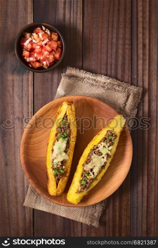 Baked ripe plantain stuffed with mincemeat, olive, green bell pepper and onion, sprinkled with cheese, a traditional dish in Central America called Canoa de Platano (Plantain Canoe), served with tomato and onion salad, photographed overhead on dark wood with natural light