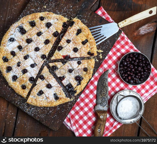 baked round black currant cake cut into pieces and sprinkled with powdered sugar on a brown wooden board