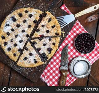 baked round black currant cake cut into pieces and sprinkled with powdered sugar on a brown wooden board