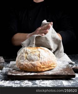 baked round bread on a board behind the cook in black clothes wipes his hands on a gray textile napkin