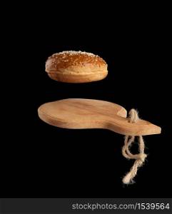 baked round bun with sesame seeds hover over brown wooden cutting board, black background, hamburger ingredient