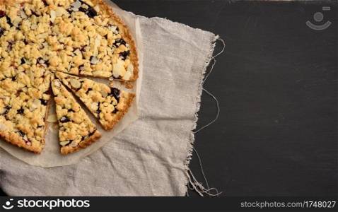 baked round crumble pie with plum cut into pieces on a black background, top view