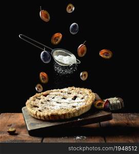 baked round pie with plums on a levitating wooden kitchen board, top of a sieve with powdered sugar and livitating slices of plum fruit on a black background