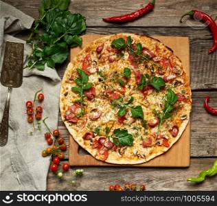 baked round pizza with smoked sausages, mushrooms, tomatoes, cheese and arugula leaves, food is cut in portions, wooden table