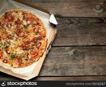 baked round pizza with smoked sausages, mushrooms, tomatoes, cheese and dill in an open cardboard box on a wooden table, food is sliced in portions, top view