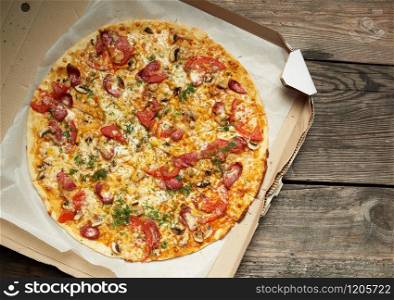 baked round pizza with smoked sausages, mushrooms, tomatoes, cheese and dill in an open cardboard box on a wooden table, food is sliced in portions, top view