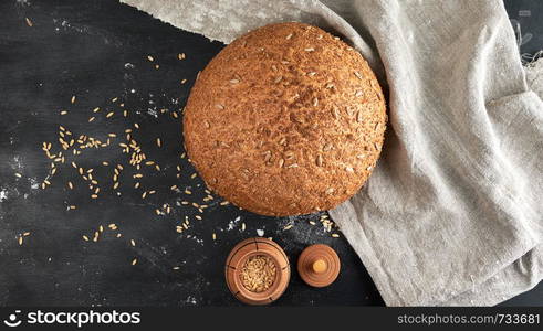 baked round rye bread with sunflower seeds on a gray textile napkin, black wooden background, top view