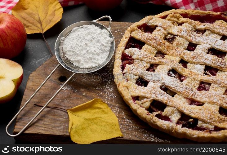 baked round traditional apple pie on a brown wooden board and fresh red apples. Next to it is an iron strainer with white powdered sugar