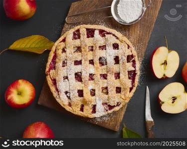 baked round traditional apple pie on brown wooden board and fresh red apples, top view