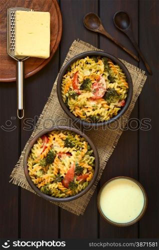 Baked tricolor fusilli pasta and vegetable (broccoli, tomato) casserole in rustic bowls, cream sauce, spoons, grater and cheese on the side, photographed overhead on dark wood with natural light