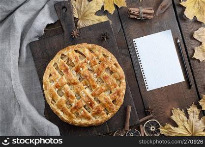 baked whole round apple pie on a brown wooden board and a blank piece of paper with a black wooden pencil, puff pastry, top view