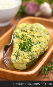 Baked zucchini stuffed with mashed potato, cheese and herbs (thyme, oregano, parsley, garlic) garnished with thyme served on wooden plate with a fork beside and a bowl of cooked rice in the back (Selective Focus, Focus on the front of the thyme garnish on the zucchini). Stuffed Baked Zucchini