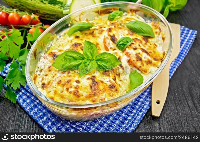 Baked zucchini with tomatoes, sour cream and cheese in a glass roaster on a blue kitchen towel, parsley and basil on wooden board background