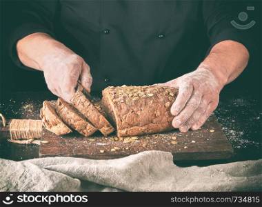 baker cuts a knife into slices of rye bread with pumpkin seeds on a brown wooden board, black background