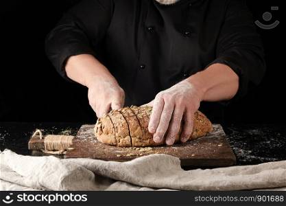 baker in black uniform cuts a knife into slices of rye bread with pumpkin seeds on a brown wooden board, black background