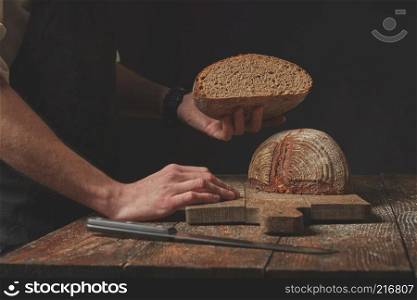 Baker keeps half freshly baked organic bread on a dark background. Hands holding half bread