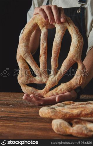Baker lays freshly baked fougas bread on a wooden brown table. Hands men holding fougas bread