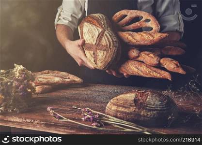 Baker with bread in hand and a wooden brown table with dry flowers on a black background, tonned photo. Variety of bread hold men&rsquo;s hands