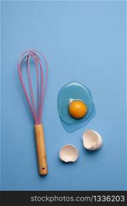 Baking and cooking concept with a kitchen utensil and broken egg. Flat lay with a cracked egg and pink whisk on a blue kitchen table.