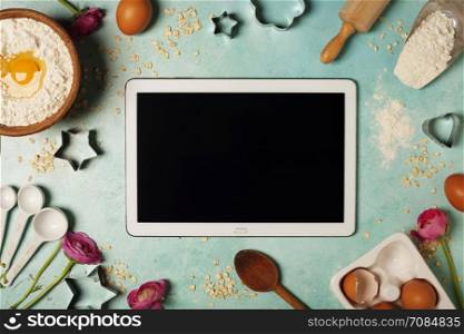 Baking background with flour, eggs, kitchen tools and tablet computer on blue rustic table. Top view. Flat lay style. Cooking meets technology