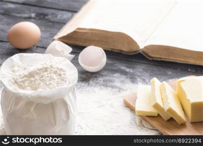 Baking theme image with a bag of wheat flour, butter and eggs, on a black wooden table, and an open book in the background.
