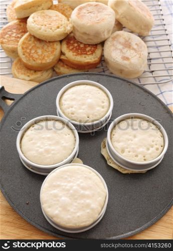Baking traditional English crumpets in rings on a hot griddle, with a pile of homemade crumpet and muffins in the background
