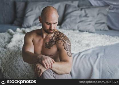 Bald beefy man with naked torso sitting in the room.. A brutal man with tattoo on shoulder and chest 1.