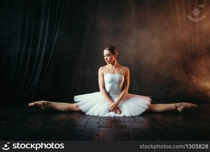 Ballerina in white dress sits on a twine, front view. Body flexibility of classical ballet dancer