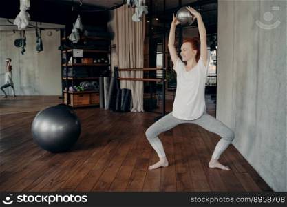 Ballet leg training. Young smiling red haired female working out in fitness studio holding small ball with both hands above head while sitting in pile with knees wide apart, looking away from camera. Sportive woman training with small pilates ball in half sitting position during ballet workout