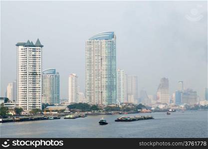 Bangkok city has a lot of tall buildings. Adjacent river boat traffic.