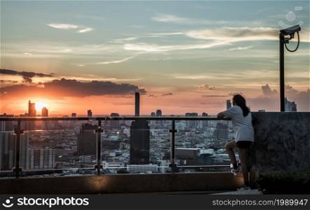 Bangkok, Thailand - 03 Jul 2021 : A young woman relaxing on rooftop terrace with overlooking the city at dawn view. New morning, Blurred town on background, Copy space, No focus, specifically.