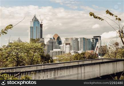 Bangkok, Thailand - Feb 19, 2022 : View of Modern high buildings and walkway bridge among surrounded green trees space against blue sky with clouds at afternoon. City growth concept, Selective focus.