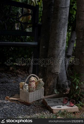 Bangkok, Thailand. May - 07, 2022 : Fresh waxy corn or Sweet glutinous corn in Wicker basket with book and sunglasses under the tree. Relaxing time on holiday at the park, Copy space, Selective focus.