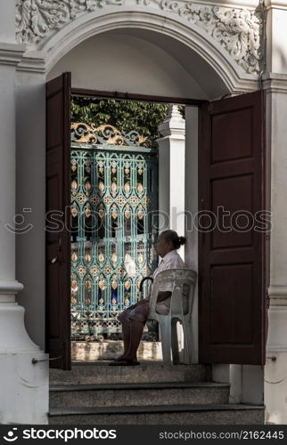 Bangkok, Thailand - Nov 15, 2020 : Elderly woman sitting in plastic chair on temple door on afternoon. No focus, specifically.