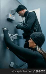 Bank robbery, male and female robbers in black uniform trying to open vault lock. Criminal profession, theft concept. Robbers in black uniform trying to open vault lock