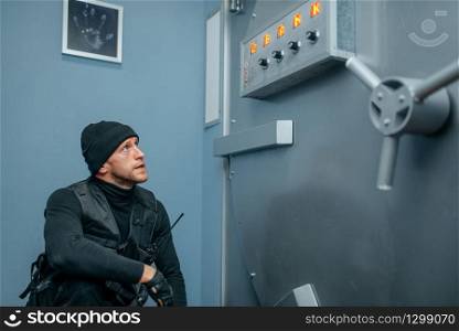 Bank robbery, male robber in black uniform sitting at the vault door. Criminal profession, theft concept. Robber in black uniform sitting at the vault door