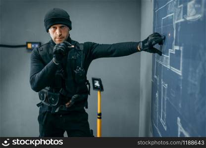 Bank robbery, male robber in black uniform, vault plan on background. Criminal profession full of risk. Theft of money and jewels concept. Bank robbery, male robber in black uniform