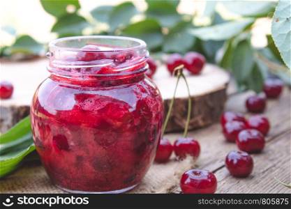 Bank with homemade cherry jam on a wooden background near the berries and leaves. Close-up.. Bank with homemade cherry jam on a wooden background near the berries and leaves.