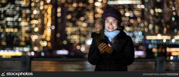Banner and cover of Asian woman in Winter suit using smart mobile phone with smile action over the photo blurred bokeh of New york Cityscape beside the east river background, USA downtown,