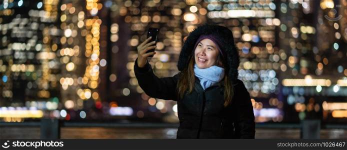 Banner and cover of Asian woman in Winter suit using smart mobile phone for selfie over the photo blurred bokeh of New york Cityscape beside the east river background, USA downtown